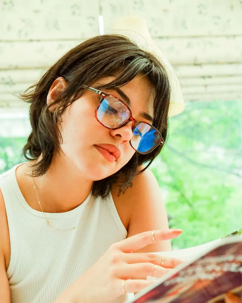 Jovem de óculos lendo uma revista perto da janela (foto ilustrativa)